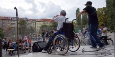Zwei Personen mit Mikrofon auf einer Bühne – eine im Rollstuhl sitzend, eine stehend – tragen etwas vor